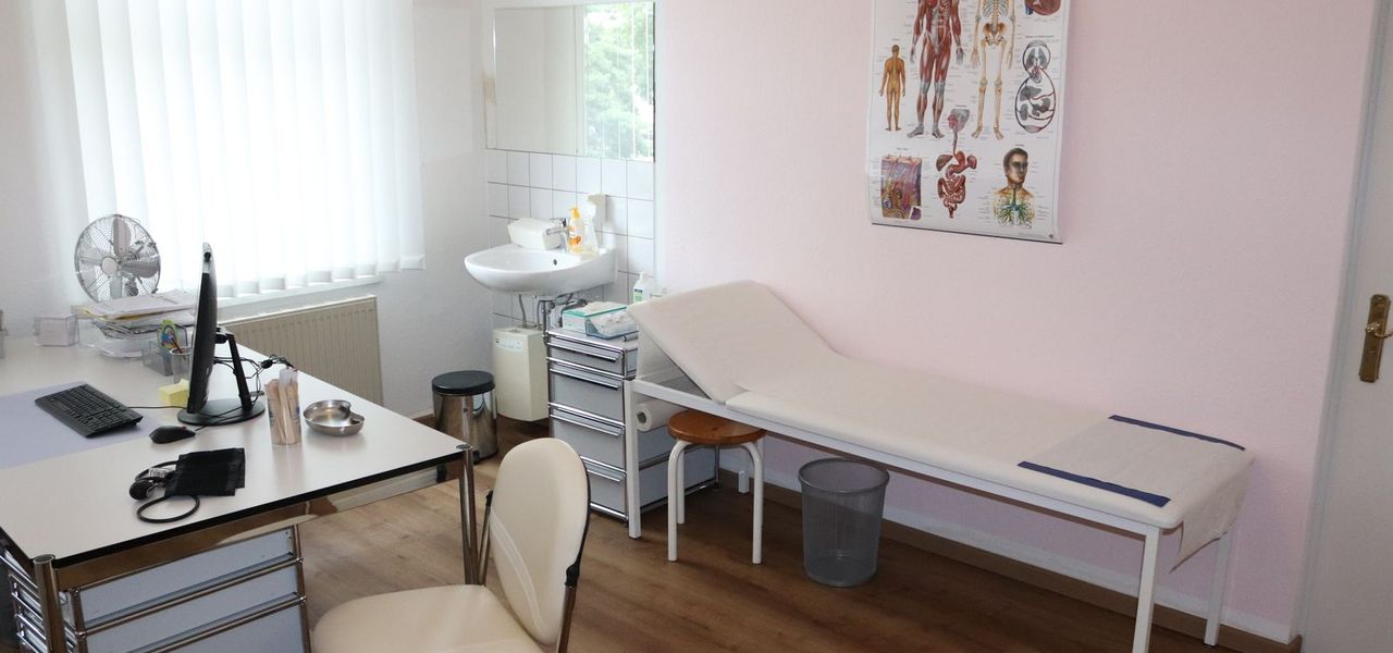 Heller Behandlungsraum mit weißer Untersuchungsliege, Schreibtisch und anatomischem Wandposter