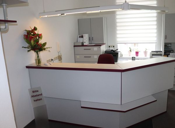 Moderner weißer Empfangstresen einer Arztpraxis mit roten Akzenten, Blumenstrauß und Hinweisschild zum Abstand halten